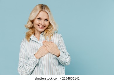 Elderly smiling kind-hearted cheerful caucasian woman 50s wear striped shirt put folded hands on heart isolated on plain pastel light blue color background studio portrait. People lifestyle concept