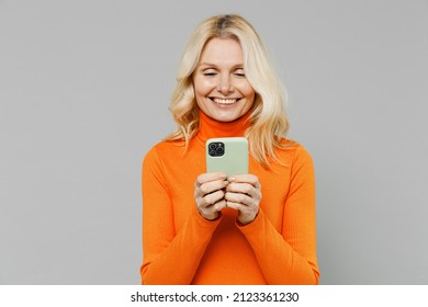 Elderly Smiling Satisfied Fun Happy Blonde Caucasian Woman 50s In Orange Turtleneck Hold In Hand Use Mobile Cell Phone Isolated On Plain Grey Color Background Studio Portrait. People Lifestyle Concept