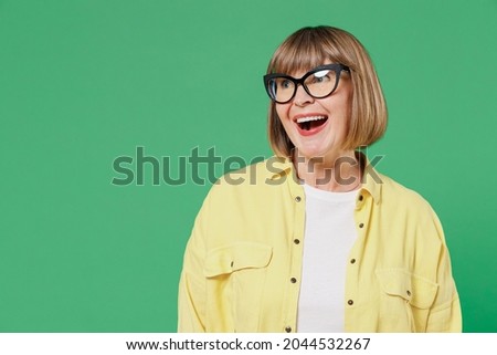 Elderly smiling surprised amazed happy caucasian woman 50s wear glasses yellow shirt look aside on workspace copy space area isolated on plain green background studio portrait People lifestyle concept
