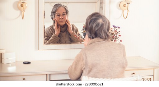 An elderly smiling woman of 80+ years of age spends a good time at home, a grandmother takes care of her appearance, looks in the mirror, sits at a dressing table in her bedroom. - Shutterstock ID 2230005521