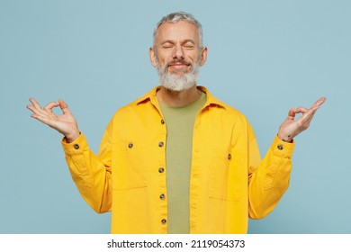 Elderly spiritual gray-haired mustache bearded man 50s wearing yellow shirt hold spreading hands in yoga om aum gesture relax meditate try to calm down isolated on plain pastel light blue background.