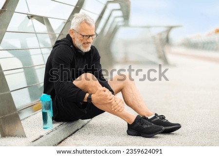 Elderly sportsman wearing black sportswear jogging outdoor by bridge, sitting on the ground and touching his leg, retired athletic man suffer from shin splint, got injured during workout, copy space