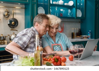 Elderly Spouses Having A Good Time Together In Kitchen. Watching Family Photos On A Laptop While Cooking A Meal.