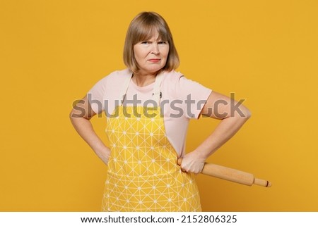 Elderly strict angry housekeeper housewife woman 50s in orange apron hold using rolling pin stand akimbo threat isolated plain on yellow background studio portrait People household lifestyle concept