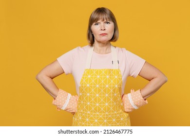 Elderly strict angry indignant sad cooker housekeeper housewife woman 50s in orange apron oven mitt stand akimbo isolated plain on yellow background studio portrait. People household lifestyle concept
