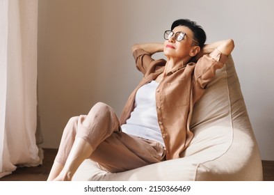 Elderly stylish woman in casual clothes and glasses holding hands behind head while resting on bean bag near window on weekend day at home