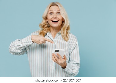 Elderly Surprised Happy Fun Woman 50s Wearing Striped Shirt Hold Use Point Finger On Mobile Cell Phone Isolated On Plain Pastel Light Blue Color Background Studio Portrait. People Lifestyle Concept