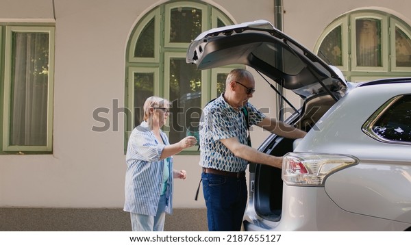 Elderly tourists getting ready for field trip\
while putting baggage and trolleys inside vehicle. Retired couple\
loading voyage luggage in car trunk while going on marriage\
anniversary summer