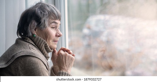 An elderly well-groomed woman, a pensioner, looks thoughtfully from the window of her house into the street. Old lady is 80+ years old with gray hair and a peaceful expression on her face.