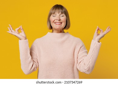Elderly woman 50s in pink casual knitted sweater hold spreading hands yoga om aum gesture relax meditate try to calm down isolated on plain yellow background studio portrait. People lifestyle concept