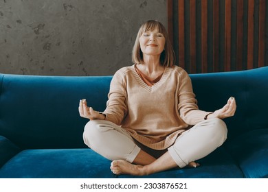 Elderly woman 50s year old wears casual clothes sit on blue sofa hold spread hands in yoga om aum gesture meditate stay at home flat rest relax spend free spare time in living room indoor grey wall - Powered by Shutterstock