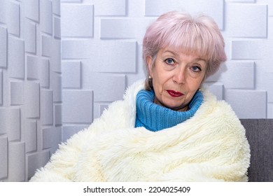 An Elderly Woman Of 60-65, Wrapped In A Warm Plaid, Warming Herself At The Heating Radiator, Looking Into The Camera. Concept: Low Indoor Temperature, Rising Gas And Heating Prices.