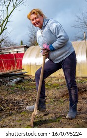 An elderly woman of 65 years old is digging in the garden against the background of a greenhouse.