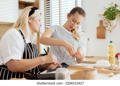 Elderly Woman In Apron Teaching Daughter Making Varennyky In Modern Kitchen With Large Window. Charming Women Having Fun Cooking Preparing Dinner Supper For Family.