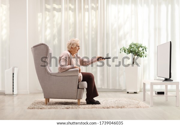 Elderly woman in an armchair changing channels on\
TV with remote control