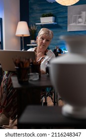 Elderly Woman Artist Living With Disability Using Laptop Looking At Vase For Inspiration In Home Art Studio. Older Person Sitting In Wheelchair Holding Portable Computer Browsing Sketch Ideas.