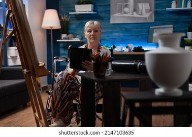 Elderly Woman Artist Living With Disability Using Tablet Computer Looking For Inspiration In Home Art Studio. Older Person Sitting In Wheelchair Holding Digital Device Browsing Sketch Ideas.