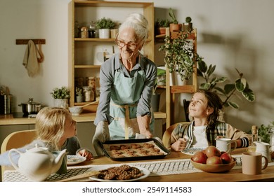 Elderly woman baking cookies with two children at kitchen table, smiling and sharing joyful moment. Kitchen is cozy with plants and wooden shelves filled with jars and utensils - Powered by Shutterstock