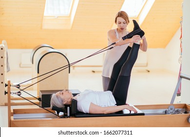 Elderly woman being helped by her instructor in the gym for exercising. Gray hair senior woman patient lying on pilates reformer raising legs up with her personal trainer assisting.