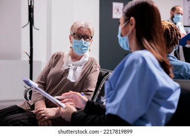 Elderly Woman Being Treated By Asian Medical Practitioner In Sanatorium. Medium Shot Of Patient At Busy Clinic, Medical Consultation. Female Doctor Interviewing Senior Female Patient At Waiting Room.