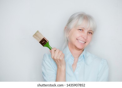 

An Elderly Woman In A Blue Shirt With A Short Sleeve And A Green Construction Tassel Is Not Against A Gray Background. Smiles And Looks Out Of A Straw Hat. Portrait. 
Looking At The Camera