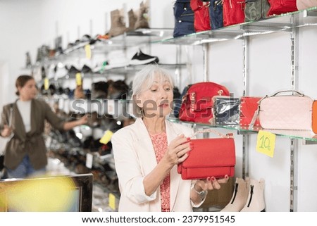 Elderly woman in casual clothes chooses leather handbag in shoe store..