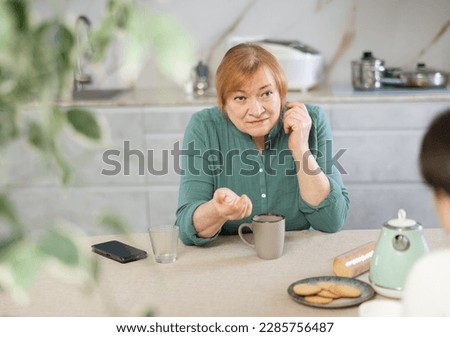 Elderly woman in casual clothes is outraged by communication with her daughter-in-law. Women talk about nasty things in kitchen at table and drink tea or coffee