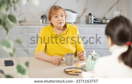 Elderly woman in casual clothes is outraged by communication with her daughter-in-law. Women talk about nasty things in kitchen at table and drink tea or coffee