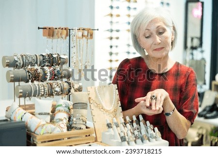 Elderly woman client enthusiastically examines showcase counter with jewelry, presenting long-awaited purchase and buying of set of semiprecious metal