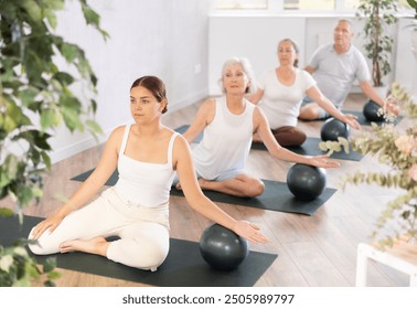 Elderly woman with company of mature friends does yoga with additional equipment. Lady is on all fours and put one palm on soft ball. Close-up - Powered by Shutterstock