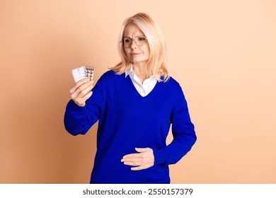 Elderly woman contemplating medication while dressed in a blue pullover against a beige background - Powered by Shutterstock