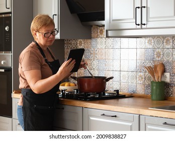 Elderly Woman Cooking In Her Kitchen And Looking For A Recipe In The Tablet. Modern Technologies And Aged People Concept