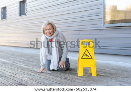 Elderly woman crawling on her knees and grimacing in pain after slipping and falling on a wet wooden deck alongside a bright yellow warning sign