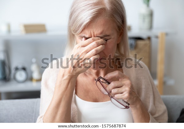 Elderly woman crying wipes tears with hands feels\
unhappy, bad news. Middle-aged woman taking off glasses closed eyes\
rubbing eyelid suffers from eye strain deterioration eyesight with\
age concept