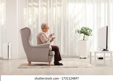 Elderly Woman With A Cup Of Coffee Seated In An Armchair In Front Of Tv At Home