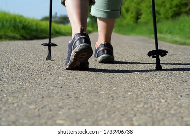 Elderly woman does Nordic Walking on an asphalted field road. Female legs with Nordic Walking poles on a cartway at a sunny day in spring.