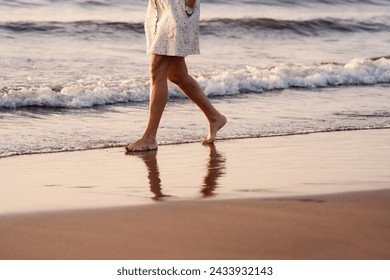 An elderly woman in a dress walks alone on the beach at sunset, reflecting on the wet sand. Senior Woman Walking Barefoot on Beach. - Powered by Shutterstock