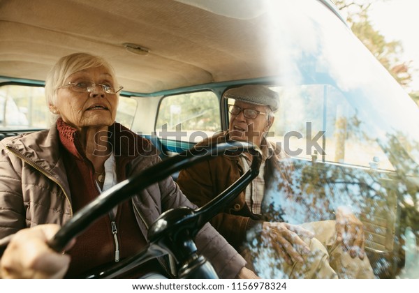Elderly woman\
driving a car with a man sitting in passenger seat looking at her.\
Senior couple traveling by\
car.
