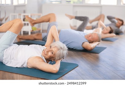 Elderly woman during group workout performs exercises to strengthen abdominal muscles, lying on back brings knee and elbow together, shakes abdominal muscles - Powered by Shutterstock