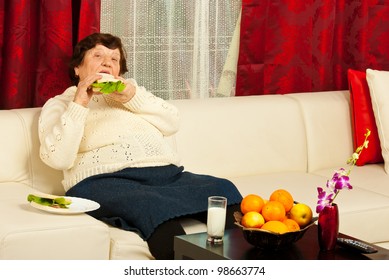 Elderly Woman Eating Sandwich And Sitting On Couch In Her Home