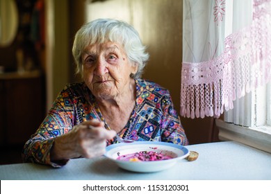 An Elderly Woman Eating Soup Sitting At A Table In The House.