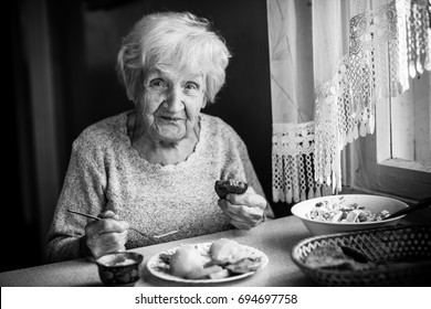 Elderly woman eats sitting at the table. Black-and-white photo.