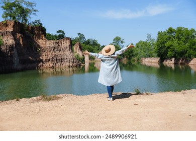 Elderly woman enjoying nature, standing with arms outstretched beside a calm pond with rocky cliffs in the background. Wearing a sun hat and casual clothing. Relishing the peaceful environment - Powered by Shutterstock