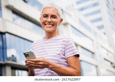 Mujer mayor que disfruta de un día soleado en la ciudad, sosteniendo su teléfono mientras que sonríe felizmente al aire libre en una camiseta casual elegante