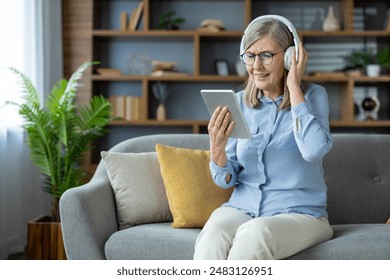 Elderly woman enjoying technology, listening to music or a podcast with headphones and using a tablet. She is sitting comfortably on a sofa in a contemporary living room setting. - Powered by Shutterstock