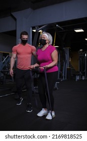 Elderly Woman Exercising With Resistance Band, Her And Personal Trainer Wearing Medical Face Mask