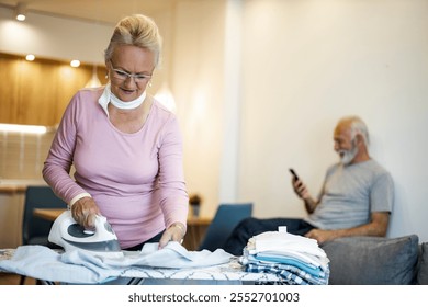 An elderly woman focuses on ironing clothes, highlighting domestic chores. In the background, an older man sits using a smartphone, creating a scene of everyday home life. - Powered by Shutterstock