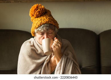 Elderly woman in ginger cap wrapped in blanket and drinking cup of tea. Old grandma feeling cold at home. Energy crisis. - Shutterstock ID 2220836443