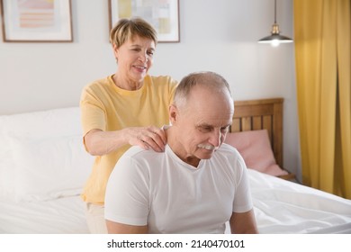 elderly woman gives a shoulder massage to her husband sitting on a bed at home. elderly couple healthy lifestyle. 