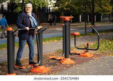 An elderly woman with glasses and gray hair engages in fitness training on outdoor exercise equipment in an urban park during autumn. Dressed in comfortable clothes, she is committed to her health - Powered by Shutterstock